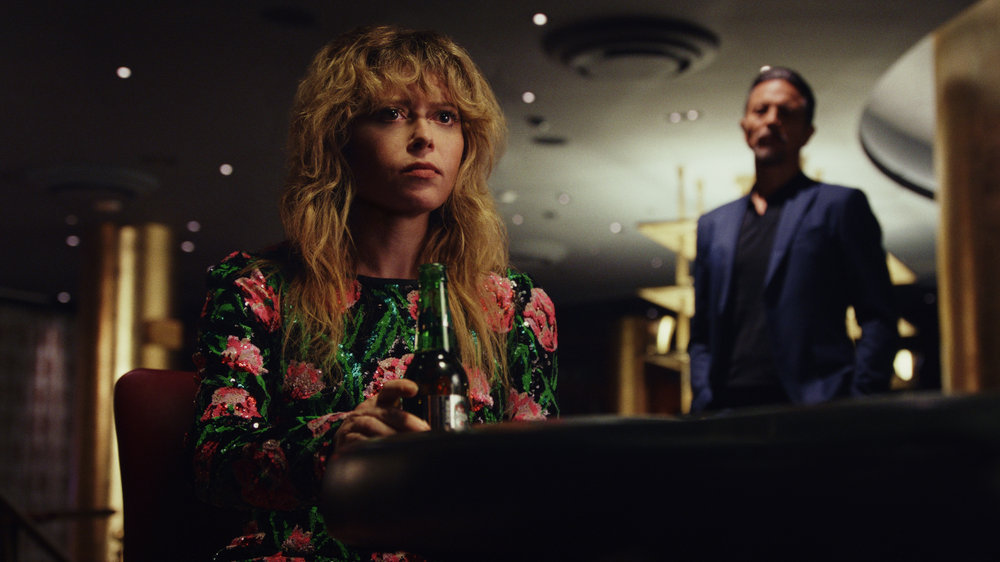 A woman in a pink and green floral sequin dress sitting at a table with a bottle of beer, while a man in a dark suit stands menacingly in the background; still from "Poker Face"