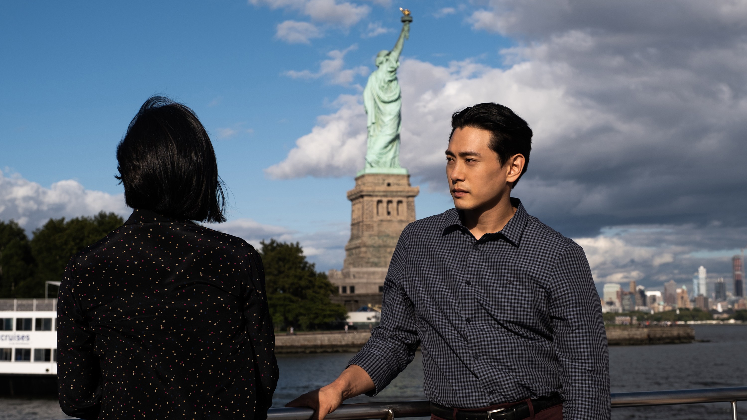 Greta Lee as Nora and Tao Yoo as Hae Sung, on a ferry sailing around the Statue of Liberty in "Past Lives." Nora is faced away from the camera looking at the statue. Hae Sung faces towards camera looking pensively at Nora.