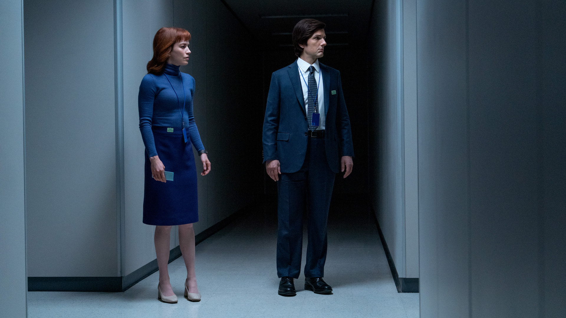 A woman in blue and a man wearing a dark suit walking in a dark white-walled hallway; still from 'Severance' Season 2