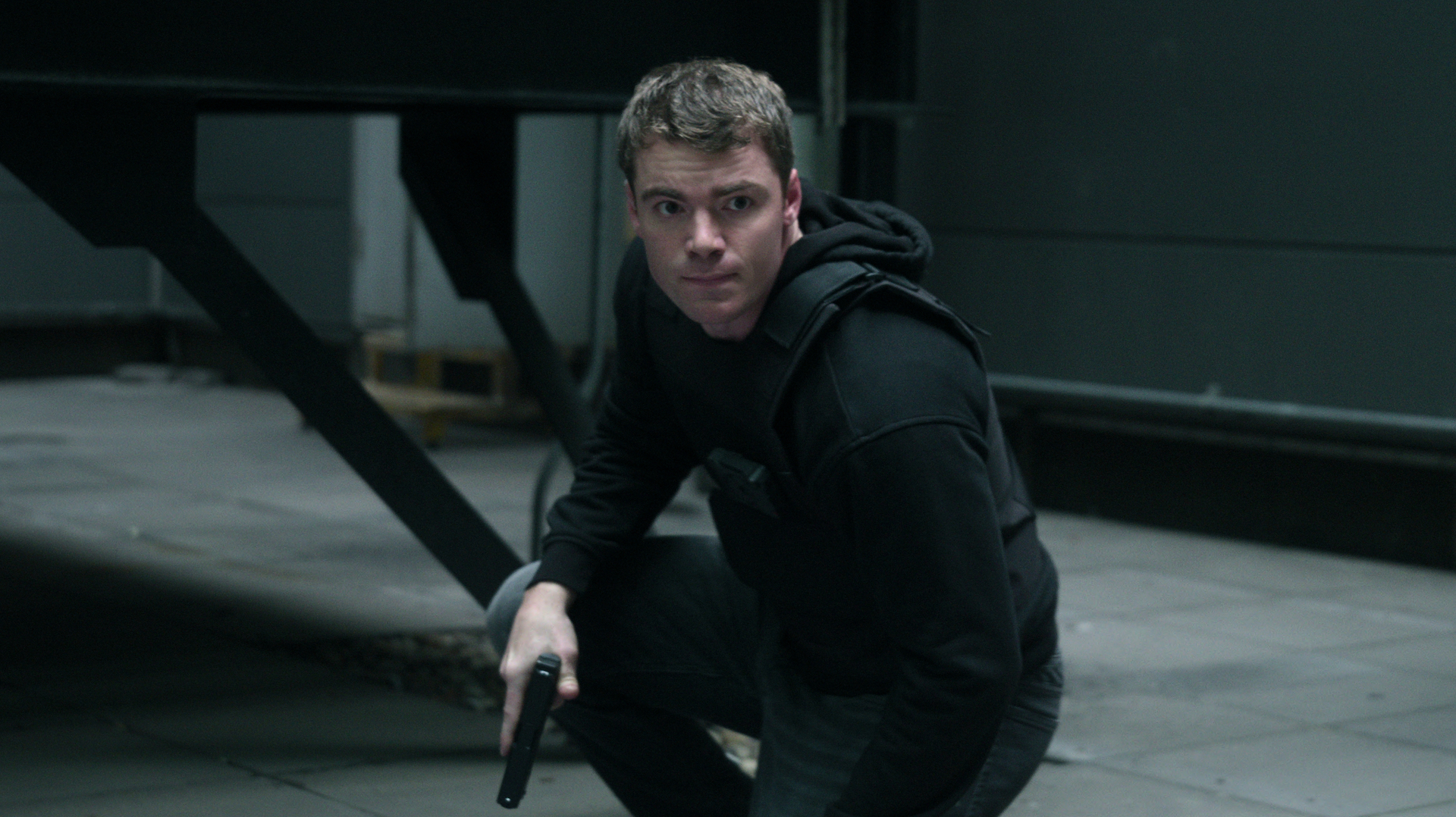 A man wearing all black and crouched while holding a handgun; Gabriel Basso in 'The Night Agent'